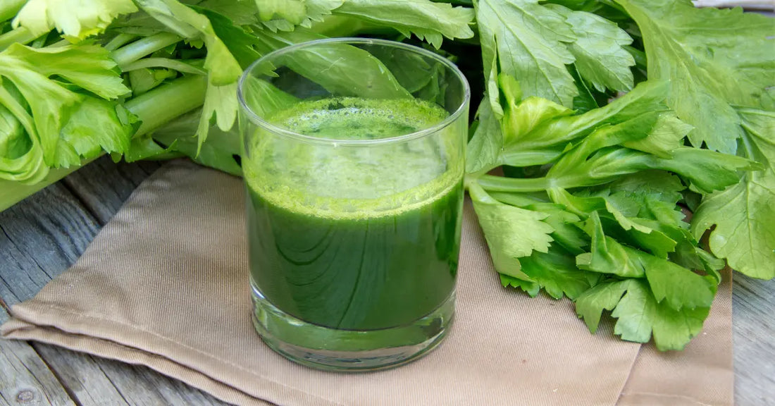 Glass of cold-pressed celery juice on a cloth napkin surrounded by leafy celery stalks