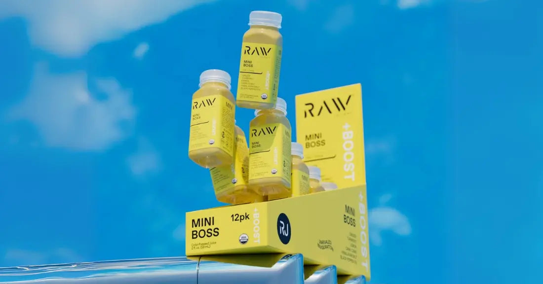 Cold-pressed turmeric juice bottles labeled "Mini Boss" stacked in a yellow display box against a bright blue sky.