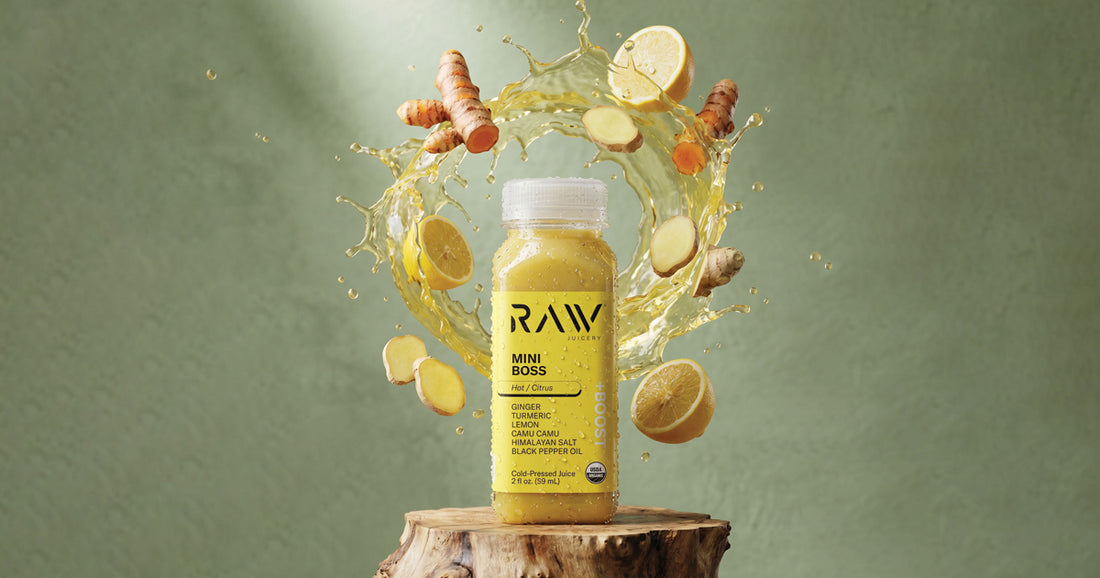 Raw Juicery Mini Boss ginger and turmeric cold-pressed shot with fresh ginger root, turmeric root, and lemon slices in a citrus splash on a wooden stump